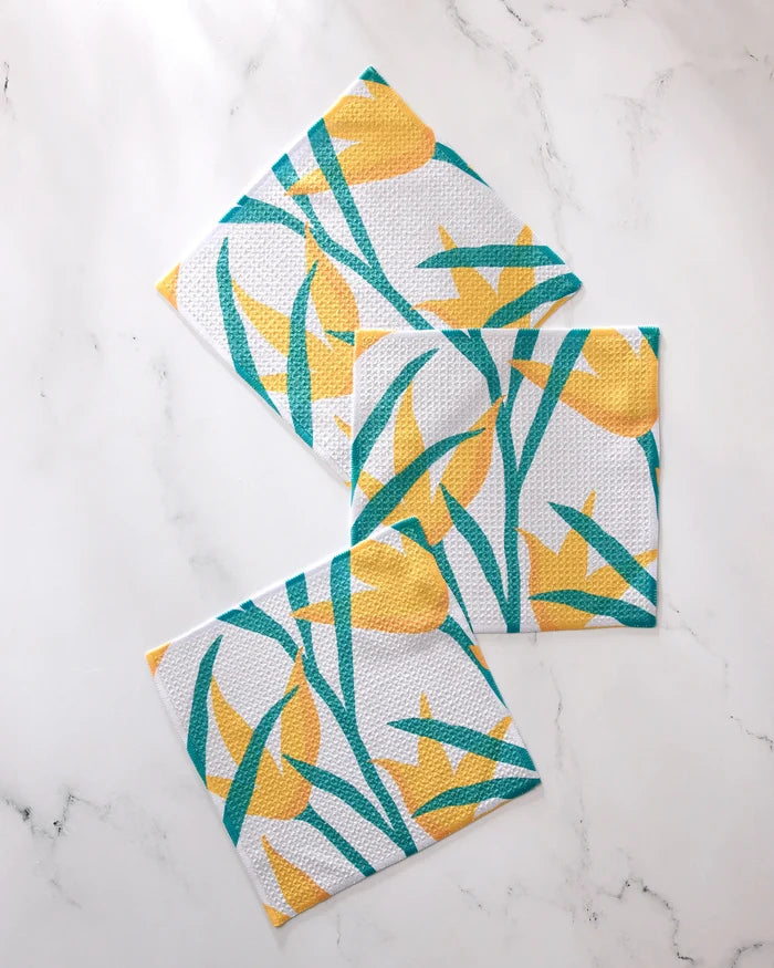 Three GEOMETRY "Growth" dishtowels with yellow flower and green leaf designs are arranged in a geometric, slightly overlapping layout on a white marble surface. This super-absorbent set brightens any kitchen.