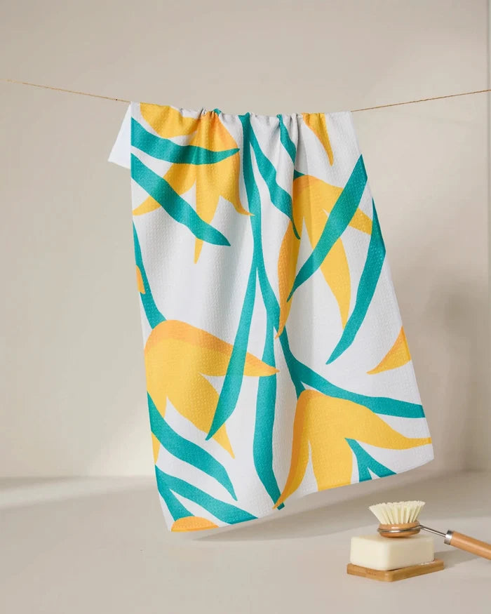 The GEOMETRY Growth tea towel, featuring a yellow and teal leaf pattern, hangs on a clothesline. Below, a wooden-handled dish brush rests on a beige surface, all set against a simple neutral background.
