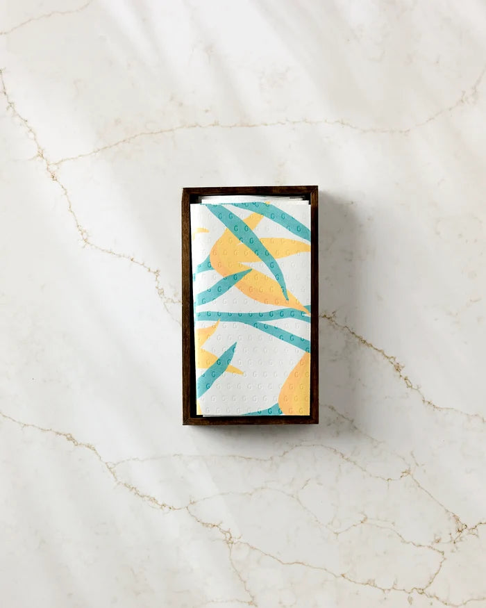 The GEOMETRY "Growth" rectangular box holds eco-friendly paper towels, with each white napkin displaying yellow and teal abstract patterns, shown atop a white marble surface with light beige veining.