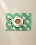 A white bowl of granola rests on the Hairy Check Green placemat by Geometry, with a spoon to the left, all set on a beige tablecloth.