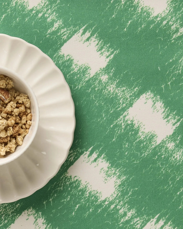 A white scalloped bowl of granola sits on Geometrys Hairy Check Green placemat, featuring a green and white brushstroke pattern and a machine-washable surface.