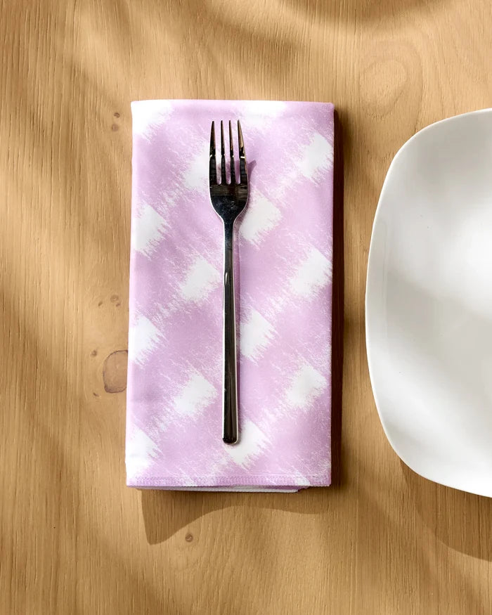 A silver fork rests on a folded Geometry Hairy Check Pink cloth napkin, placed on a light wooden table next to the edge of a white square plate.