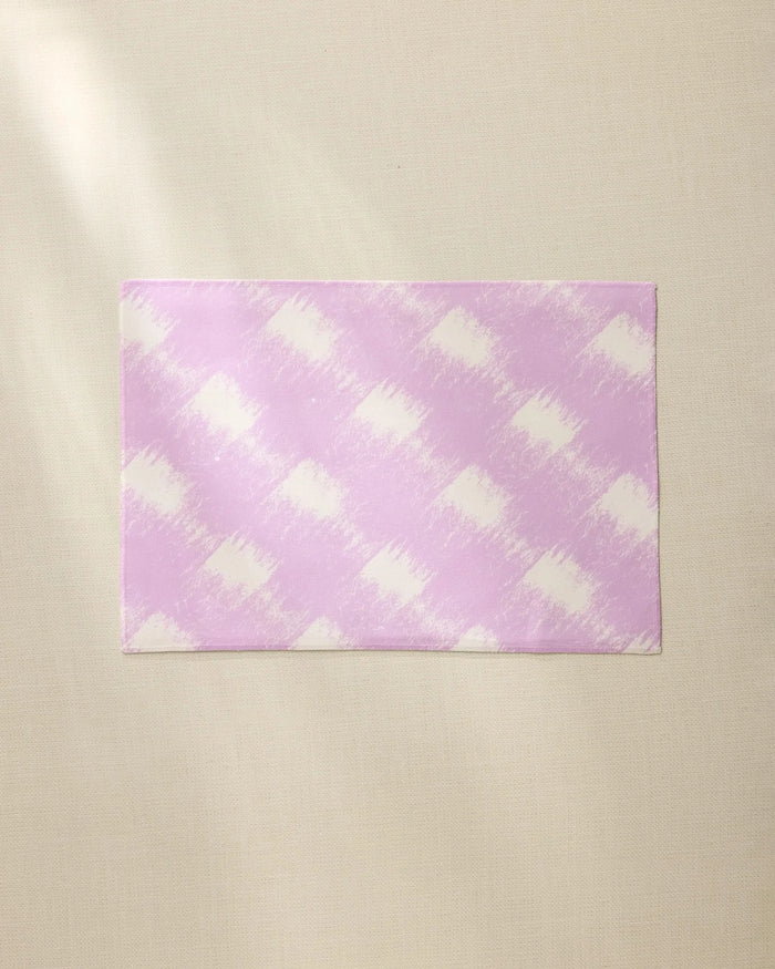 The Geometry Hairy Check Pink placemat features a light purple and white diagonal checkered pattern. Durable and machine washable, its a stylish, practical choice for everyday dining. Shown laid flat on a beige textured surface.