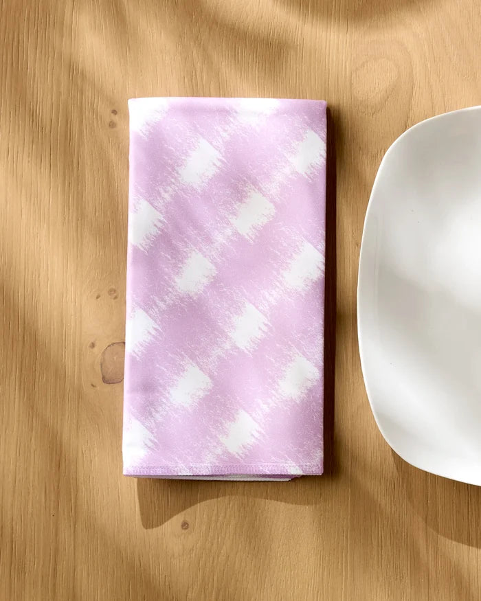 A folded Geometry Hairy Check Pink napkin from the Dinner Napkin Set rests on a light wooden table beside the edge of a plain white plate.