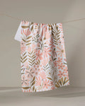 The "Halia" recycled kitchen towel by Amy MacCready, featuring pink and gold floral and leaf patterns, hangs on a clothesline against a plain beige background.