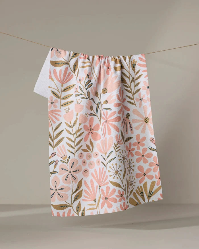 The "Halia" recycled kitchen towel by Amy MacCready, featuring pink and gold floral and leaf patterns, hangs on a clothesline against a plain beige background.