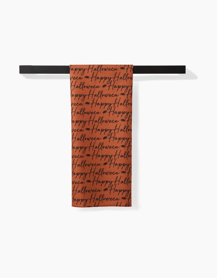 The Geometry Halloween Script Orange tea towel features a festive Happy Halloween black script on an orange background, hanging on a black bar. This ultra-absorbent towel brings seasonal charm and practicality to your kitchen.