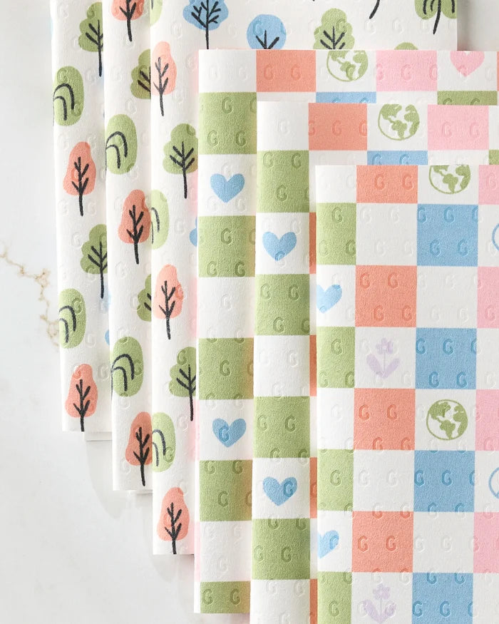 Kayla Ann’s Happy Checkers set includes four eco-friendly kitchen towels: one with pastel blue-green trees, and three with checkered hearts, flowers, and globes in pink, purple, blue, and green on a white background.
