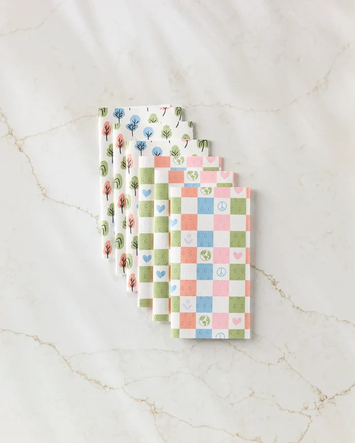 Three folded Kayla Ann Happy Checkers eco-friendly kitchen towels with pastel tree, peace, heart, and earth motifs plus pink, blue, and green checked designs are arranged on a marble surface.