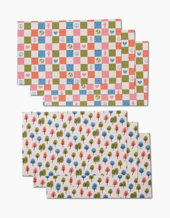 Two sets of Kayla Ann Happy Checkers placemats: one with a pink, green, red, and blue checkered design featuring peace signs, hearts, and smiley faces; the other with rows of small trees. Both are made from environmentally friendly materials.