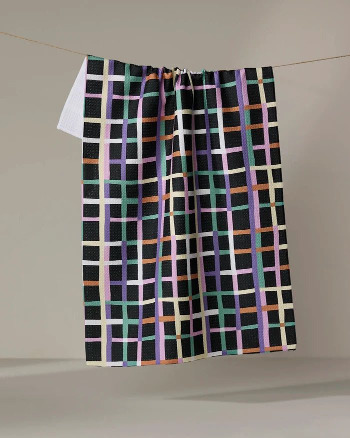 The Happy Plaid tea towel by Claire Iglesias features a vibrant green, lavender, peach, white, and orange grid on black—ultra-absorbent and perfect for brightening any kitchen with its colorful design.