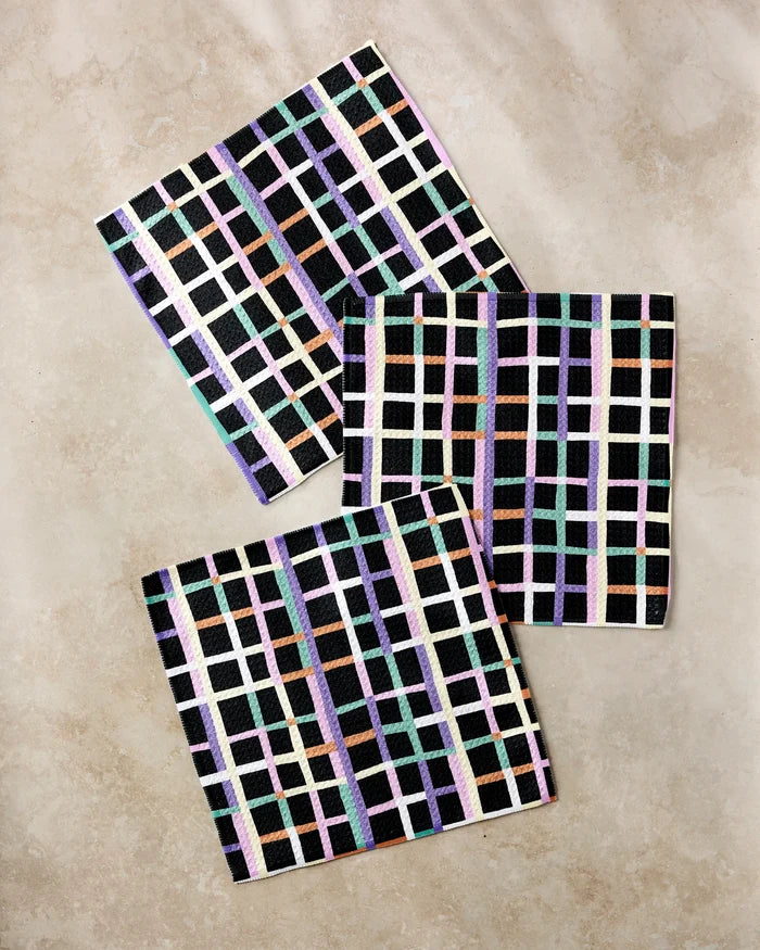 The Happy Plaid napkins by Claire Iglesias are eco-friendly, square fabric napkins made from recycled materials, featuring a vibrant pastel grid on black. These stylish napkins add charm to any dishcloth set.