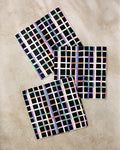 The Happy Plaid napkins by Claire Iglesias are eco-friendly, square fabric napkins made from recycled materials, featuring a vibrant pastel grid on black. These stylish napkins add charm to any dishcloth set.