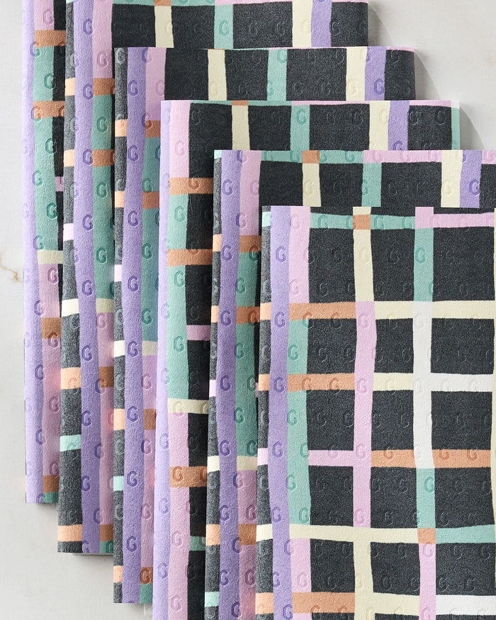 Eight folded Happy Plaid reusable paper towels by Claire Iglesias—featuring black, white, mint green, lavender, peach, and yellow pastel checks—are fanned out on a light marble surface.