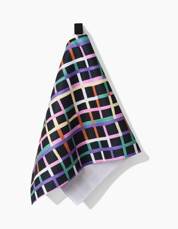 The Happy Plaid Bar Towel by Claire Iglesias features a vibrant grid of purple, pink, green, orange, and white lines on black Geoweave fabric and hangs from a black loop against a white background.