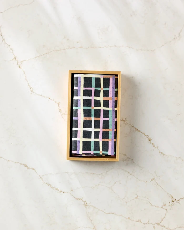 A rectangular box of Claire Iglesias's Happy Plaid reusable paper towels, featuring an abstract pastel grid pattern and a black item inside, rests on a light marbled surface.