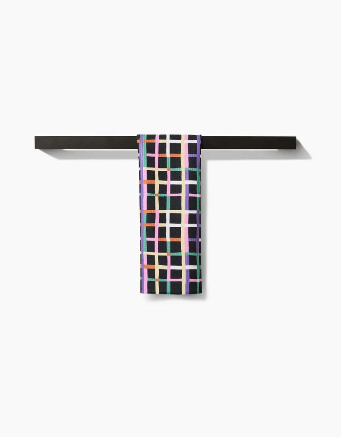 The Happy Plaid bar towel by Claire Iglesias, made from soft Geoweave fabric and featuring a colorful geometric grid design, hangs neatly over a minimalist black towel bar against a plain white background.