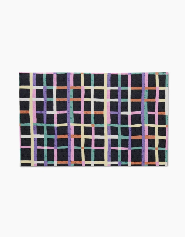 The Happy Plaid rug by Claire Iglesias features a grid pattern of intersecting purple, teal, beige, orange, and green lines on a dark background. Made from post-consumer recycled materials, it’s an eco-friendly choice for your space.