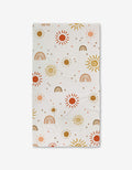 The GEOMETRY Happy Skies Luxe Bath Towel features a cheerful pattern of suns, rainbows, and dots in brown, yellow, red, and pink on ultra-absorbent fabric evenly spread across the white surface.