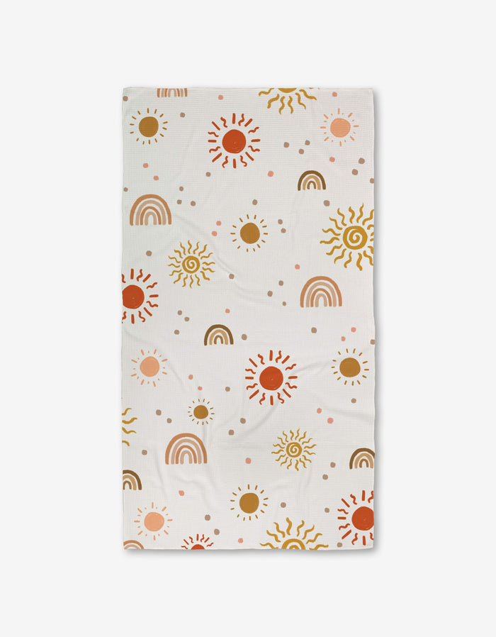 The GEOMETRY Happy Skies Luxe Bath Towel features a cheerful pattern of suns, rainbows, and dots in brown, yellow, red, and pink on ultra-absorbent fabric evenly spread across the white surface.