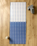 The Harmony Haze by Happy Hemera, a non-slip yoga towel with a blue and white checkered pattern, is spread on a wooden floor. A water bottle sits bottom left, with two gray yoga blocks at the top right of the mat.
