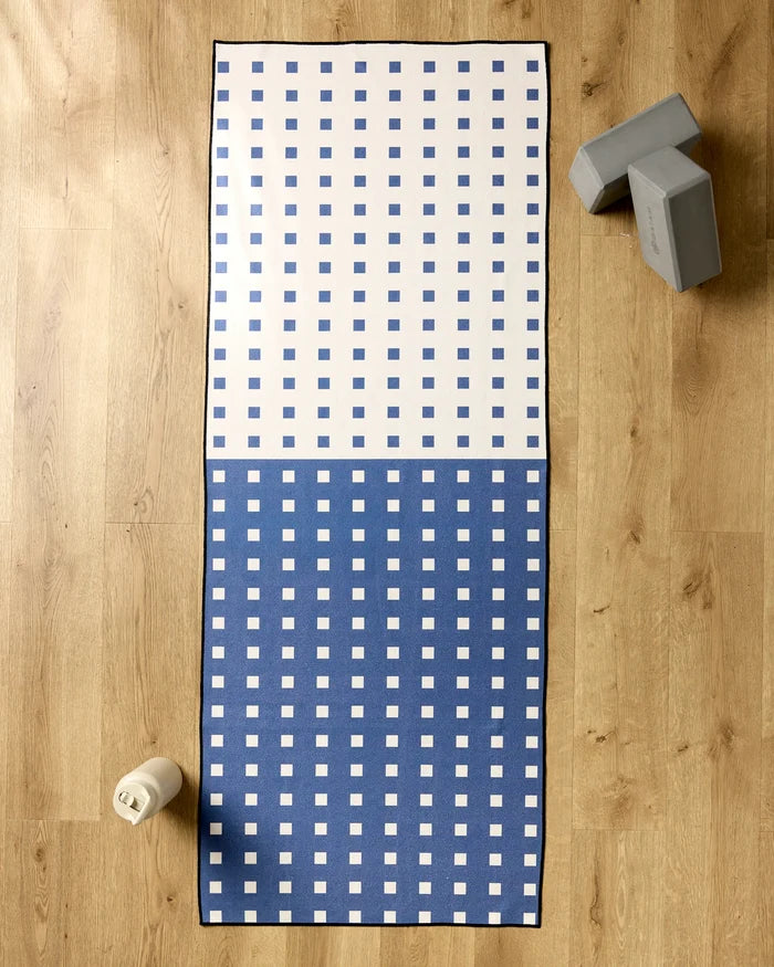 The Harmony Haze by Happy Hemera, a non-slip yoga towel with a blue and white checkered pattern, is spread on a wooden floor. A water bottle sits bottom left, with two gray yoga blocks at the top right of the mat.