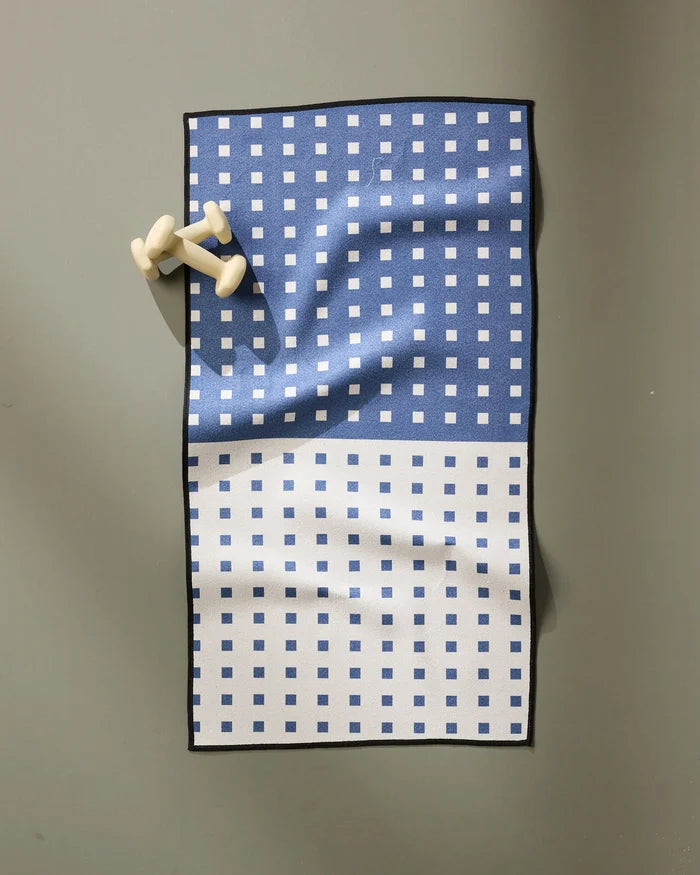 The Harmony Haze fitness towel by Happy Hemera, made from recycled materials, features a blue and white checkered design and two white chess pawns near the top left corner on a gray surface.