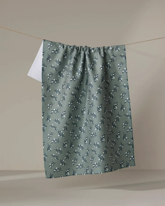The "Harvest Berries" recycled kitchen towel by Charlie Rowan Designs, featuring a green leaf and berry pattern, hangs on a clothesline with its white reverse side showing against a plain beige background.