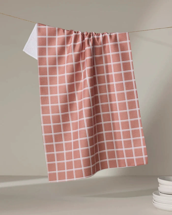 The Harvest Plaid tea towel by Charlie Rowan Designs hangs on a string against a neutral background, with a stack of white plates partially visible in the lower right corner.