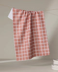 The Harvest Plaid tea towel by Charlie Rowan Designs hangs on a string against a neutral background, with a stack of white plates partially visible in the lower right corner.