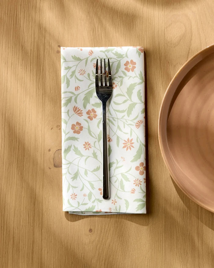 A silver fork rests on Danelys Sidron's Hawthorns, an eco-friendly table linen with green and orange floral patterns, laid on a light wooden table beside a peach-colored plate.