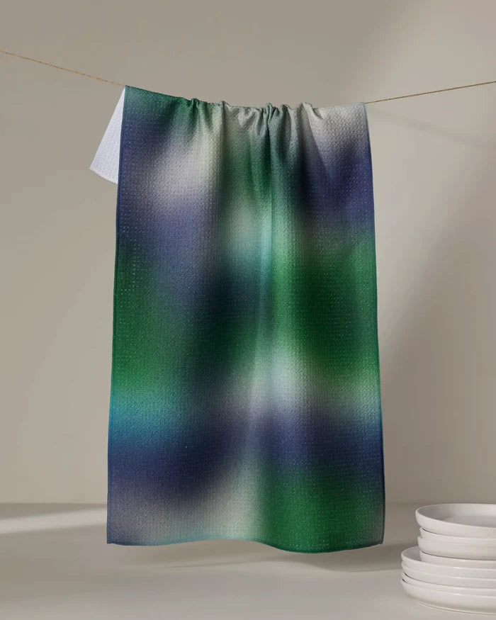 The Geometry Haze dish towel, featuring a green, blue, and white gradient pattern, hangs on a clothesline above a stack of white dishes against a neutral background.