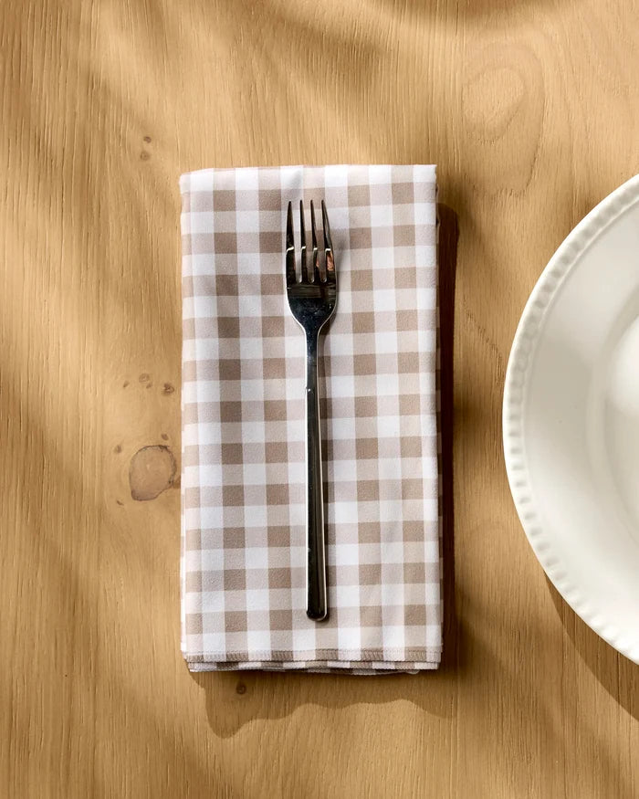 A metal fork rests on the Geometry Hazel Gingham beige and white checkered dinner napkin set, placed on a light wood table beside a white plate as sunlight casts gentle shadows across the scene.