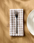 A metal fork rests on the Geometry Hazel Gingham beige and white checkered dinner napkin set, placed on a light wood table beside a white plate as sunlight casts gentle shadows across the scene.