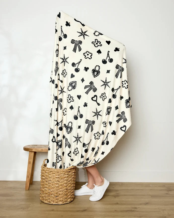 A person stands in a woven basket, mostly covered by Geometry's silky Heart Locket blanket with black abstract designs. Only their legs and white sneakers are visible. Beside them, a wooden stool rests against a plain wall.