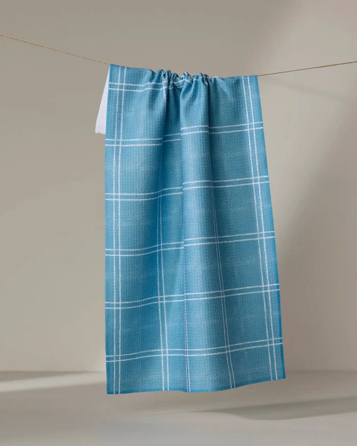 The Geometry Hearth Bay kitchen towel, featuring a blue recycled fabric with a white plaid pattern, hangs on a string against a neutral background.