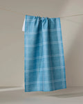 The Geometry Hearth Bay kitchen towel, featuring a blue recycled fabric with a white plaid pattern, hangs on a string against a neutral background.
