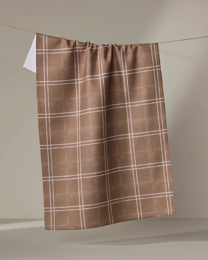 The Geometry Hearth Chestnut tea towel, featuring a white checkered pattern on brown and made from recycled materials, hangs on a string against a plain beige background.