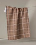 The Geometry Hearth Chestnut tea towel, featuring a white checkered pattern on brown and made from recycled materials, hangs on a string against a plain beige background.