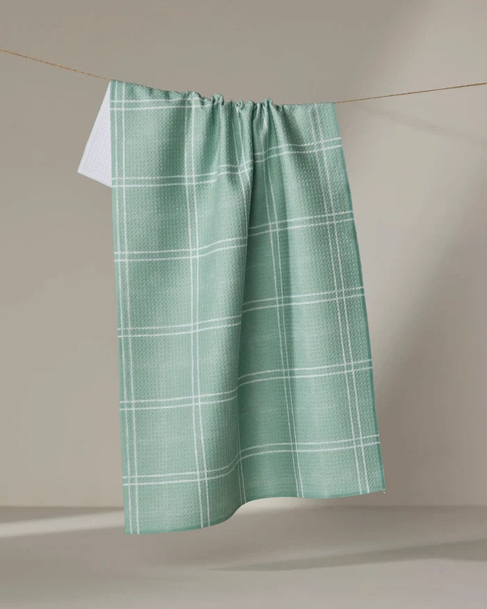 The Geometry Hearth Meadow dish towel, light green with a white checkered pattern, hangs on a clothesline against a neutral background.