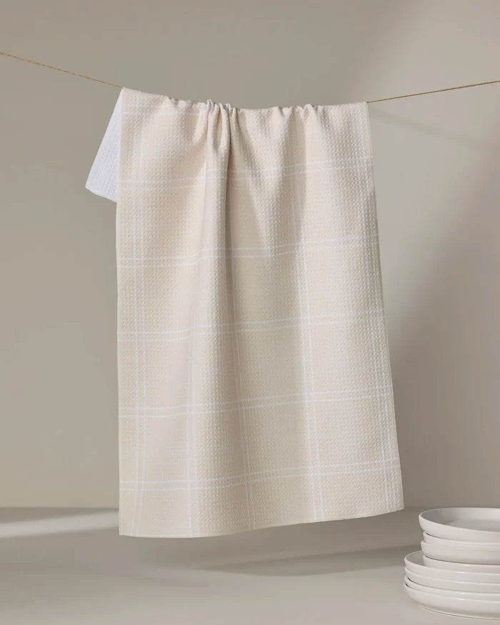 The Geometry Hearth Oat eco-friendly kitchen towel, featuring a beige and white checkered design, hangs on a string against a light background with a stack of white plates nearby.