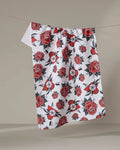 The Cole Strem "Hearts and Roses" white recycled kitchen towel, featuring a bold red and black floral pattern, hangs from a thin string against a plain beige background.