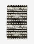 The Danelys Sidron Heartstring tea towel is rectangular and beige, made from recycled materials with black stripes, dots, hearts, and triangles in a repeating pattern. Slightly wrinkled, it is displayed on a white background.