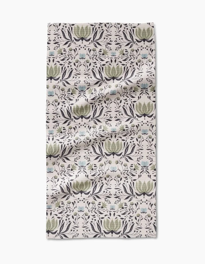 The Hello Flowers Bar Towel by Amita Nair is crafted from ultra-absorbent Geoweave fabric, featuring a green, blue, and black floral pattern on light beige. Rectangular with light wrinkles, its shown on a white surface.
