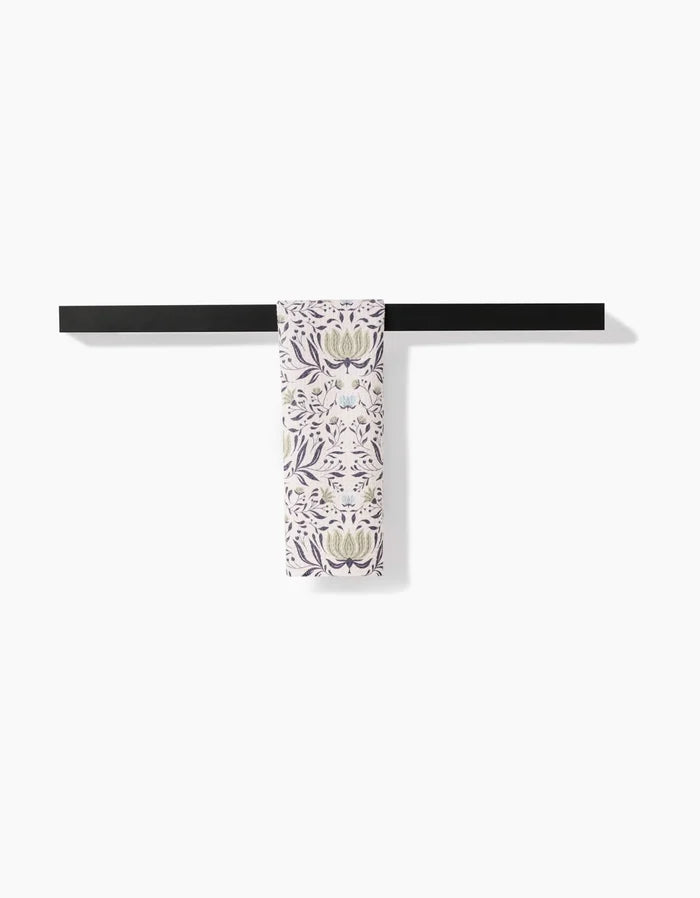 The Hello Flowers bar towel by Amita Nair, featuring a purple and green floral pattern on ultra-absorbent Geoweave fabric, is folded and draped over a black bar against a white background.