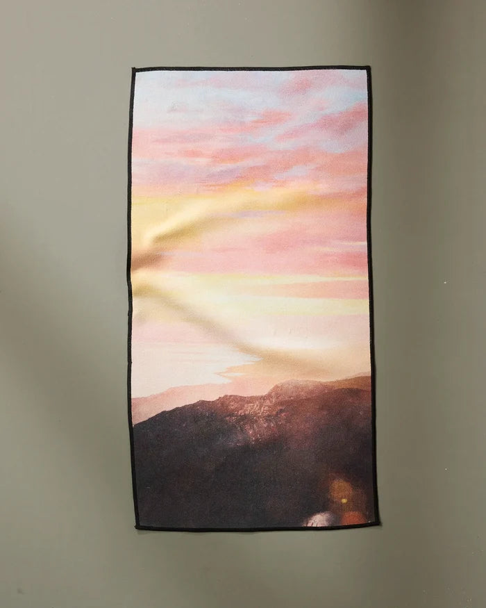 The "Her Majesty" fitness towel by Stefanie Bales is rectangular, made from recycled materials, and displays a mountain scene at sunset with pink and orange skies above shadowy hills, shown flat on a plain light gray surface.