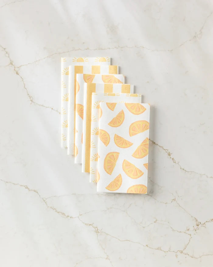 Six folded Aubrey Fairchild Here Comes The Sun white napkins, featuring yellow citrus slice patterns, are neatly arranged on a light marble surface—perfect eco-friendly kitchen towels for stylish, sustainable dining.