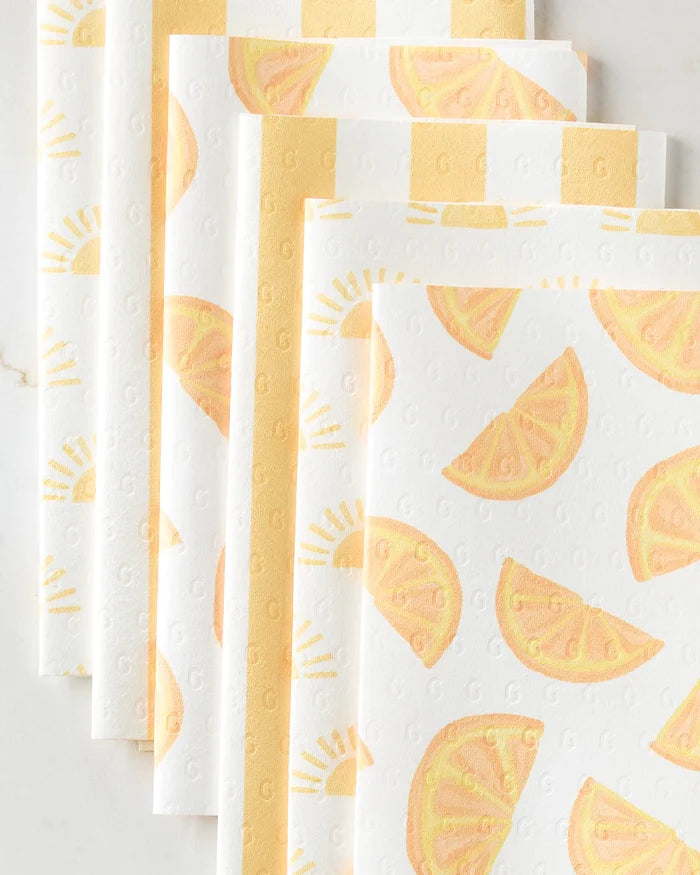 Five "Here Comes The Sun" paper napkins by Aubrey Fairchild, featuring yellow citrus slice and sun designs, are arranged in a fan pattern. Their textured surface has small repeating letter G imprints for a unique look.
