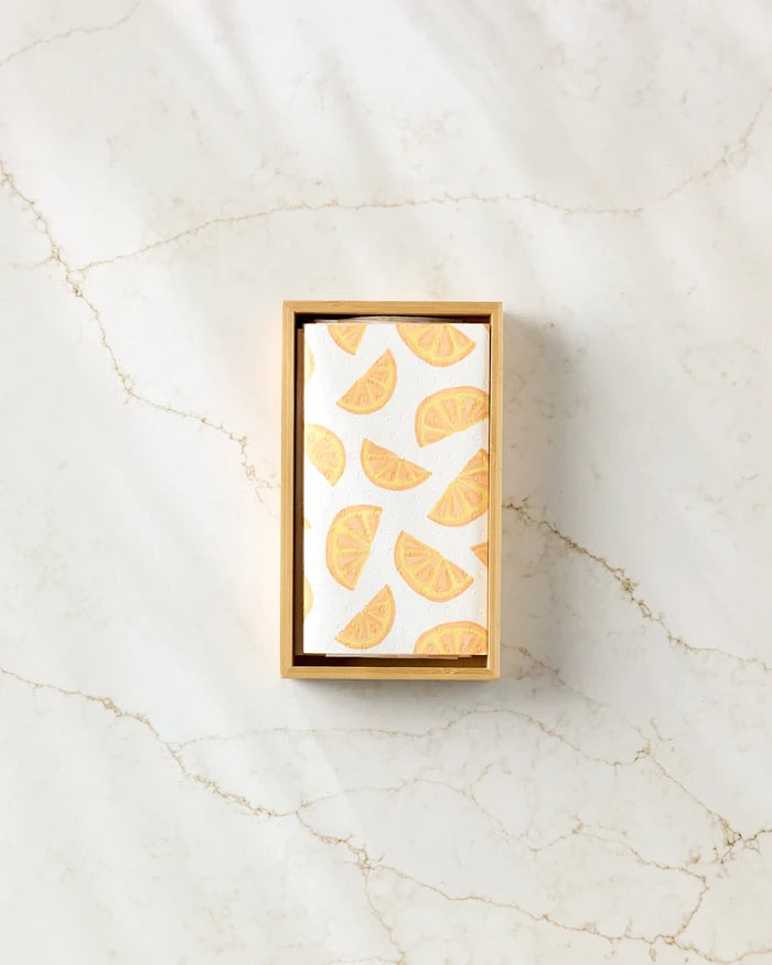 Aubrey Fairchild’s "Here Comes The Sun" eco-friendly kitchen towels, packaged in a rectangular box with a white card and orange citrus slice pattern, rest on subtly veined light marble.