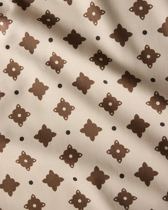 The Heritage Stamp tablecloth by Geometry features beige fabric with a repeating dark brown geometric pattern and dots, accented by subtle diagonal shadows—an eco-friendly choice perfect for modern homes.